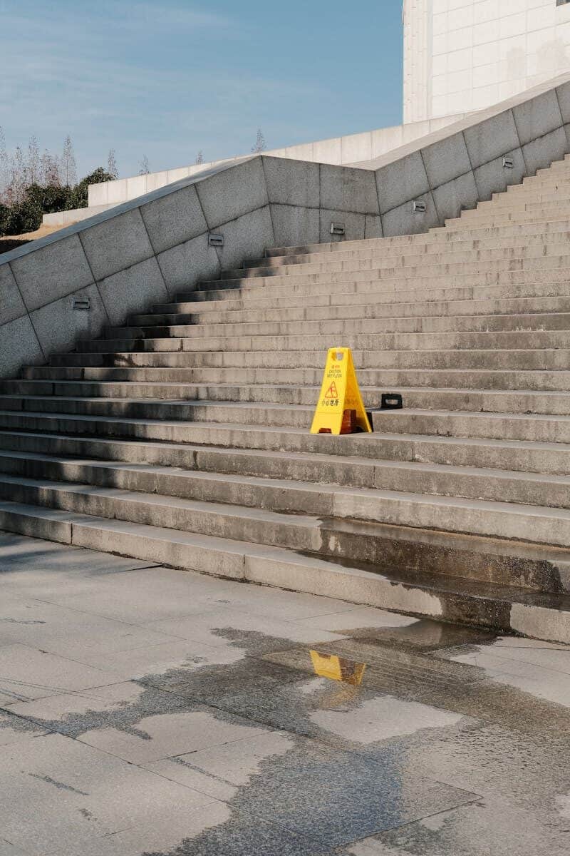 Concrete steps in an urban area with a wet floor warning sign and puddle reflection.