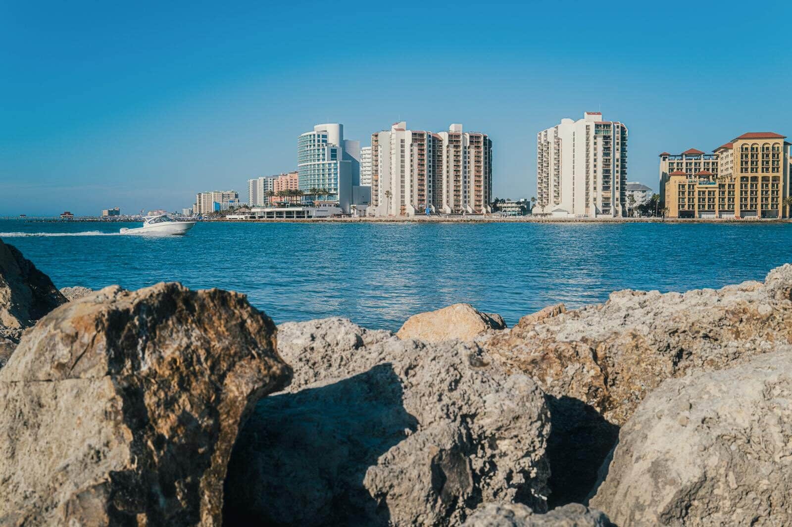 A scenic view of high-rise buildings by the water with rocks in the foreground.